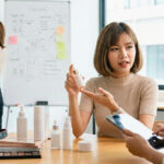 Two women discussing skincare products and content creation strategy at a desk with a professional camera, illustrating non-service beauty business roles.