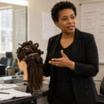 Experienced beauty instructor teaching beside a mannequin head in a cosmetology classroom with sectioning clips, lesson notes, salon tools, mirrors, and student chairs.
