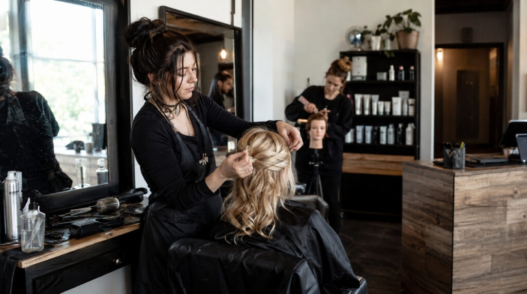 Cosmetology student completes a client hairstyle at a working salon station with appointment technology, inventory storage, and reception operations visible nearby.
