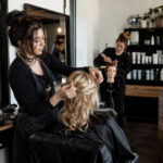 Cosmetology student completes a client hairstyle at a working salon station with appointment technology, inventory storage, and reception operations visible nearby.