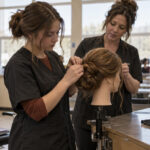 Teen student practices securing a simple mannequin updo while an adult cosmetology instructor demonstrates hairpin placement.
