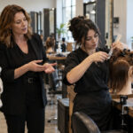 A prospective student and instructor observe a beauty student practicing a haircut on a mannequin beside a classroom workstation.